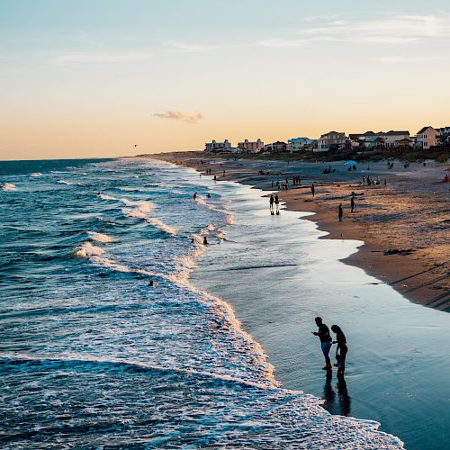 People are enjoying a beach at sunset with gentle waves and houses in the background.