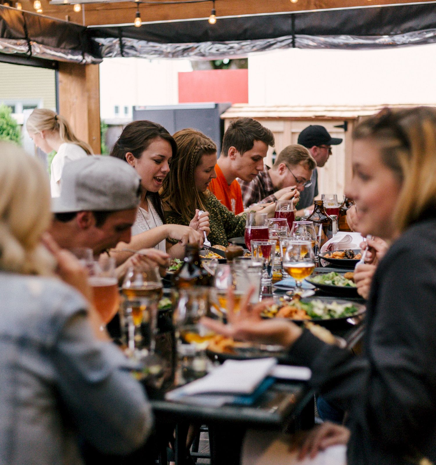 A group of people is gathered around a long table, enjoying a meal and drinks at an outdoor setting, engaged in lively conversation and laughter.