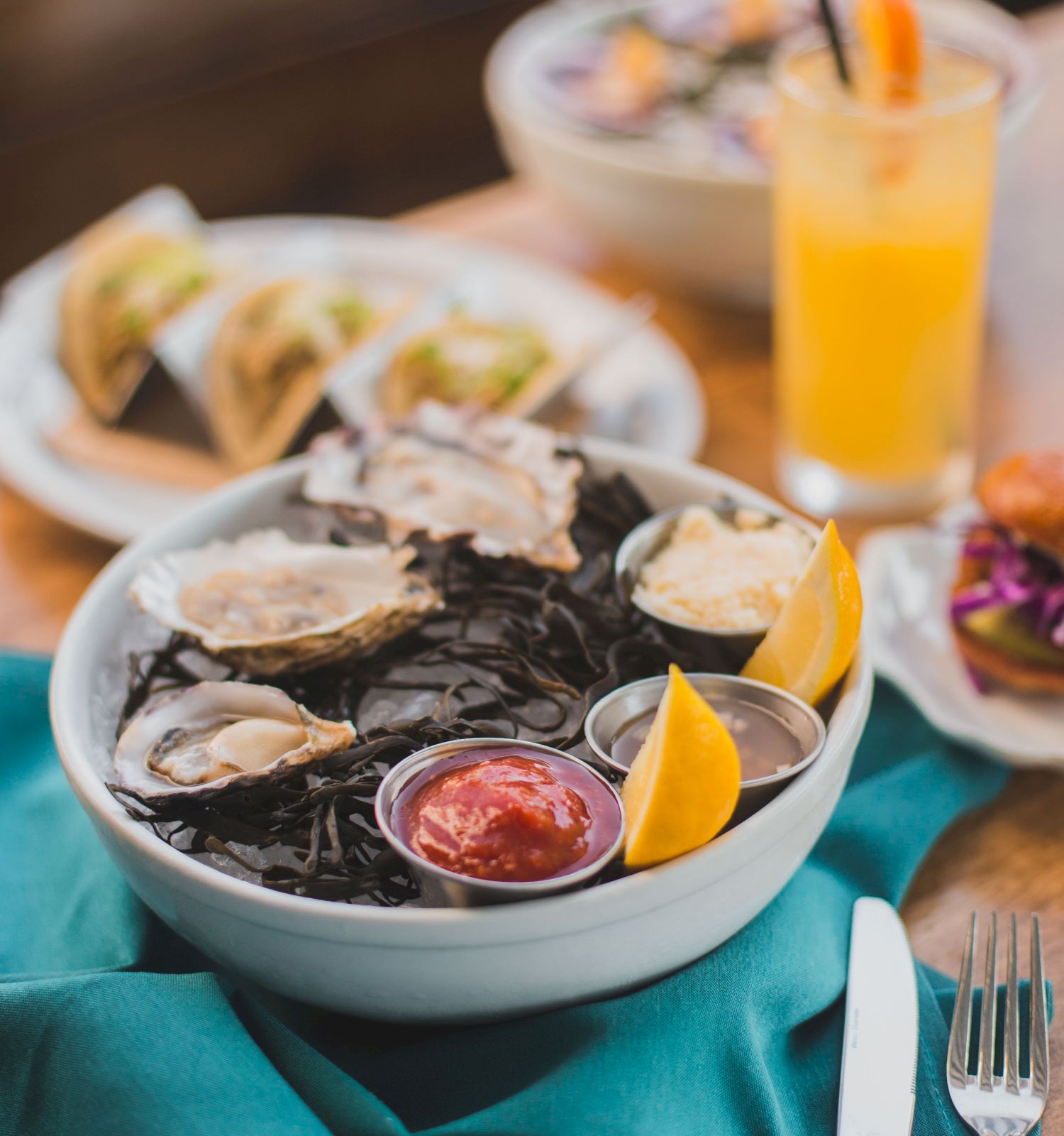 A seafood dish with oysters, lemon wedges, and sauce, a drink, and tacos in the background.