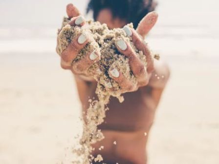 A person sitting on a beach holds up sand, letting it fall through their fingers, with an out-of-focus ocean in the background.