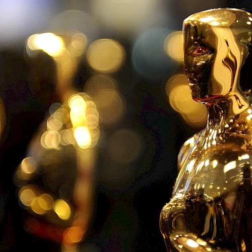 The image shows a close-up of gold Oscar statuettes, which are awarded at the Academy Awards, with a blurred background of more statues.