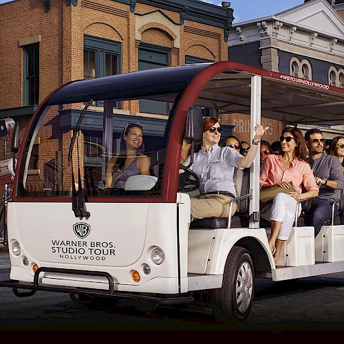 A group of people is seated in an open trolley labeled "Warner Bros. Studio Tour Hollywood" in front of studio buildings.