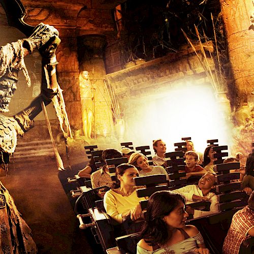 A group of people are seated on a ride or attraction with a large skeleton figure wielding a weapon above them, creating a spooky atmosphere.