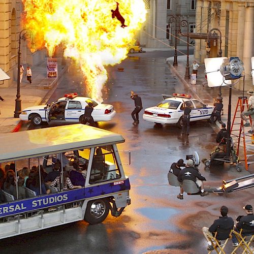 A street movie set features an explosion, police cars, actors, a film crew with equipment, and a Universal Studios tour bus with spectators.