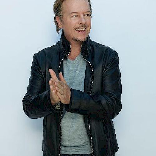 A man with light hair is wearing a grey shirt and a black leather jacket, clapping his hands and smiling against a plain white background.